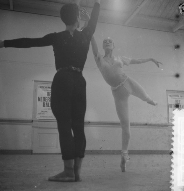 Ein Schwarz-Weiß-Foto von einem Mann und einer Frau, die in einem Studio tanzen, wobei die Frau in der Luft ist und eine plakatbedeckte Wand im Hintergrund zu sehen ist, die von oben beleuchtet wird.