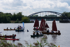 Eine Gruppe von Menschen in kleinen Booten auf einem Gewässer mit einer Brücke, Fahrzeugen, Bäumen und einem bewölkten Himmel im Hintergrund sowie Pflanzen am Boden, was eine Regatta vermuten lässt.