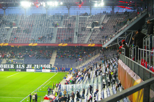 Großes Stadion voller Zuschauer bei einem Fußballspiel mit sitzenden und stehenden Personen, einem Geländer, einer rechten Banner und einem leeren Hintergrund mit Deckenbeleuchtung.