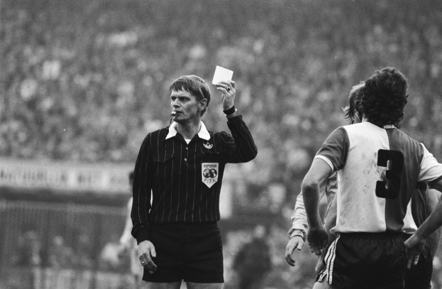 Ein Schiedsrichter in einem Schwarz-Weiß-Bild hält eine rote Karte vor einer Menge von Menschen in Fußballkleidung, mit einem Zaun im Hintergrund.