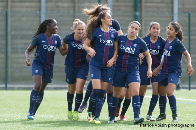 Eine Gruppe junger Frauen, die auf einem Rasenfußballfeld mit Maschendrahtzaun und einer Wand im Hintergrund spielen, mit dem Text "Paris Saint-Germain Frauenfußball" in der rechten unteren Ecke.