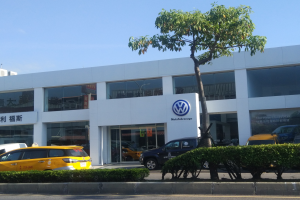 Ein Volkswagen-Autohaus mit parkenden Autos vorne, umgeben von Pflanzen und einem Baum, mit einem gläsernen Gebäude und einem sichtbaren Himmel im Hintergrund.
